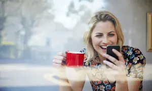 Uśmiechnięta kobieta z czerwoną filiżanką w dłoni, przegląda coś zabawnego na telefonie. Tłumacz ze zdjęcia.