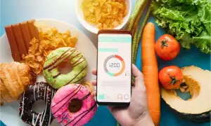 Ręka trzyma telefon z aplikacją do liczenia kalorii, obok słodkości i warzywa.
