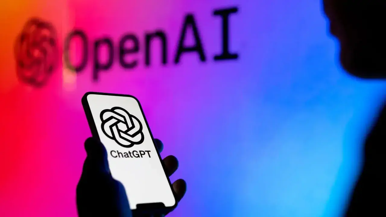 Dłoń trzyma smartfon z logo ChatGPT. W tle logo OpenAI, symbolizujące rozwój gpt sztuczna inteligencja.