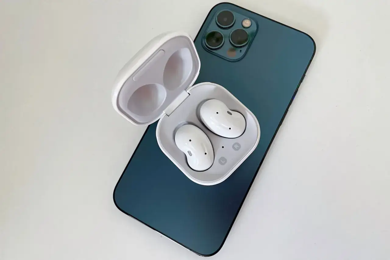 Białe słuchawki Galaxy Buds w etui leżą na granatowym iPhonie.