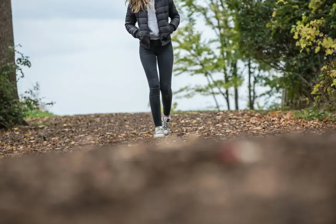 Kobieta w kurtce i jeansach idzie leśną ścieżką. Widać, co daje spacer: kontakt z naturą i świeże powietrze.