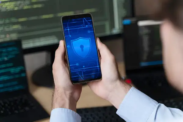 Ręce trzymają smartfon z ikoną tarczy i klucza, symbolizującą bezpieczeństwo. To wskazówka, jak odblokować telefon bez utraty danych.